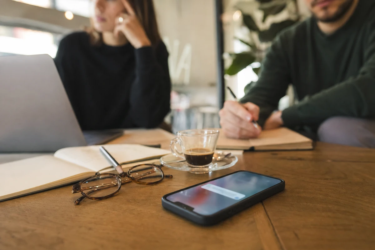 Smartphone a faccia in su su un tavolo con notifica sfocata, tazzina di caffè, occhiali e quaderno, persone sullo sfondo