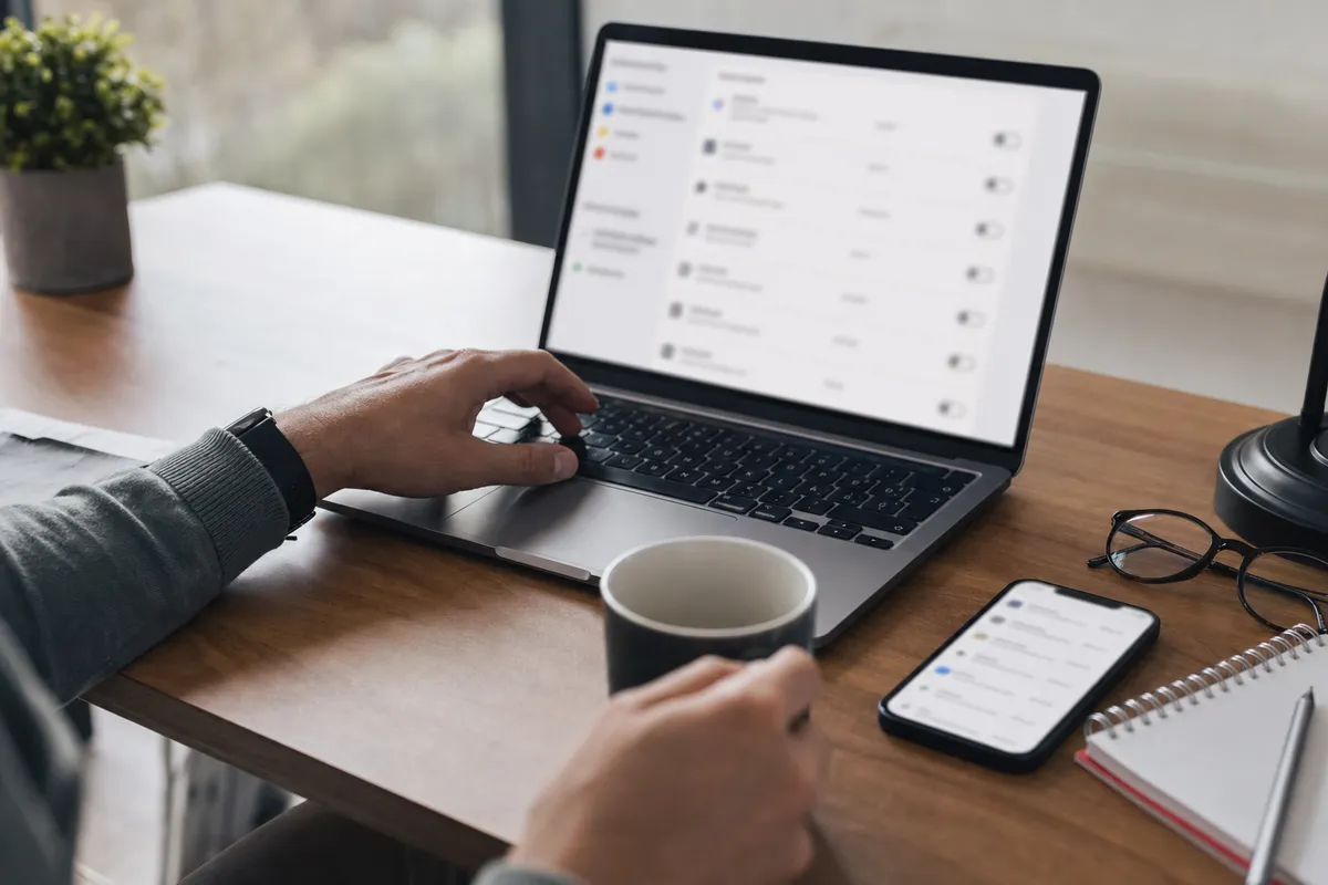 Mani su laptop con schermata sfocata di impostazioni account, smartphone sul tavolo e tazza di caffè