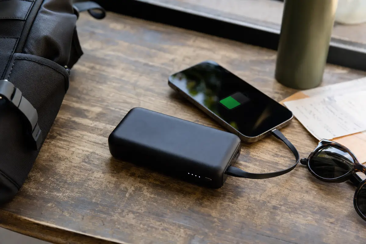 Power bank nera che ricarica uno smartphone su tavolo in legno, con zaino, occhiali da sole e borraccia sullo sfondo