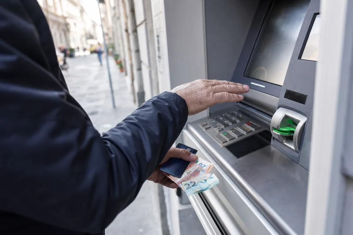 Mano che copre la tastiera di un bancomat mentre l’altra tiene carta e banconote in euro