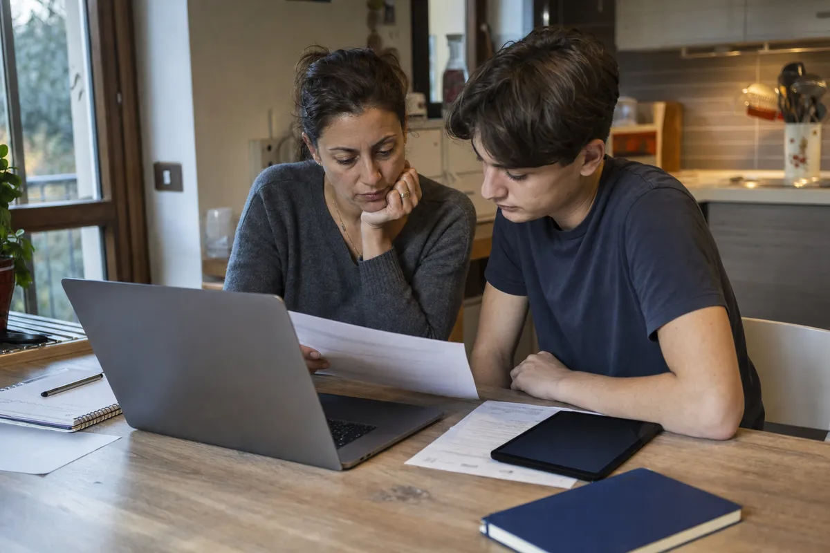 Genitore e adolescente a un tavolo in cucina con laptop aperto, documenti e tablet mentre controllano carte