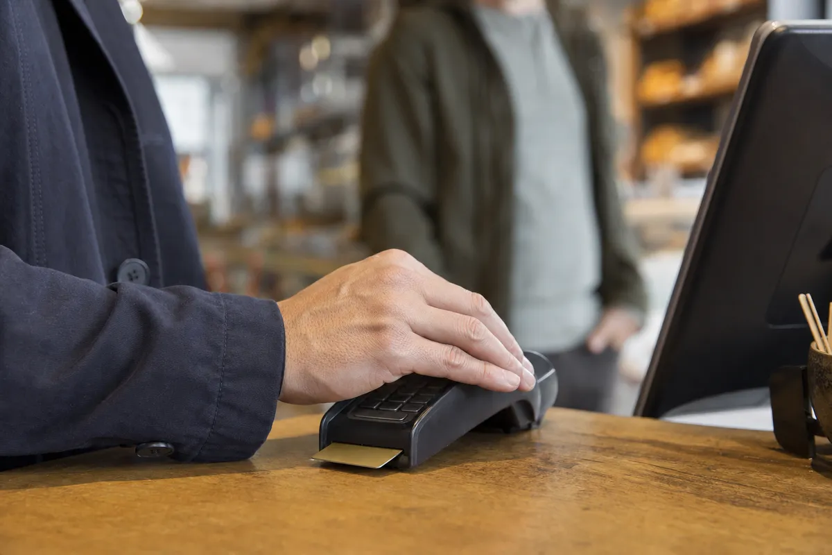 Mano che copre la tastiera del POS mentre inserisce il PIN, con carta inserita e persona sfocata sullo sfondo