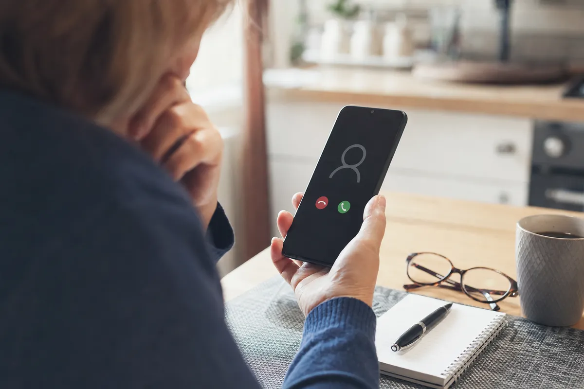 Persona a un tavolo in cucina guarda una chiamata in arrivo sullo smartphone, con tazza di caffè, occhiali e taccuino
