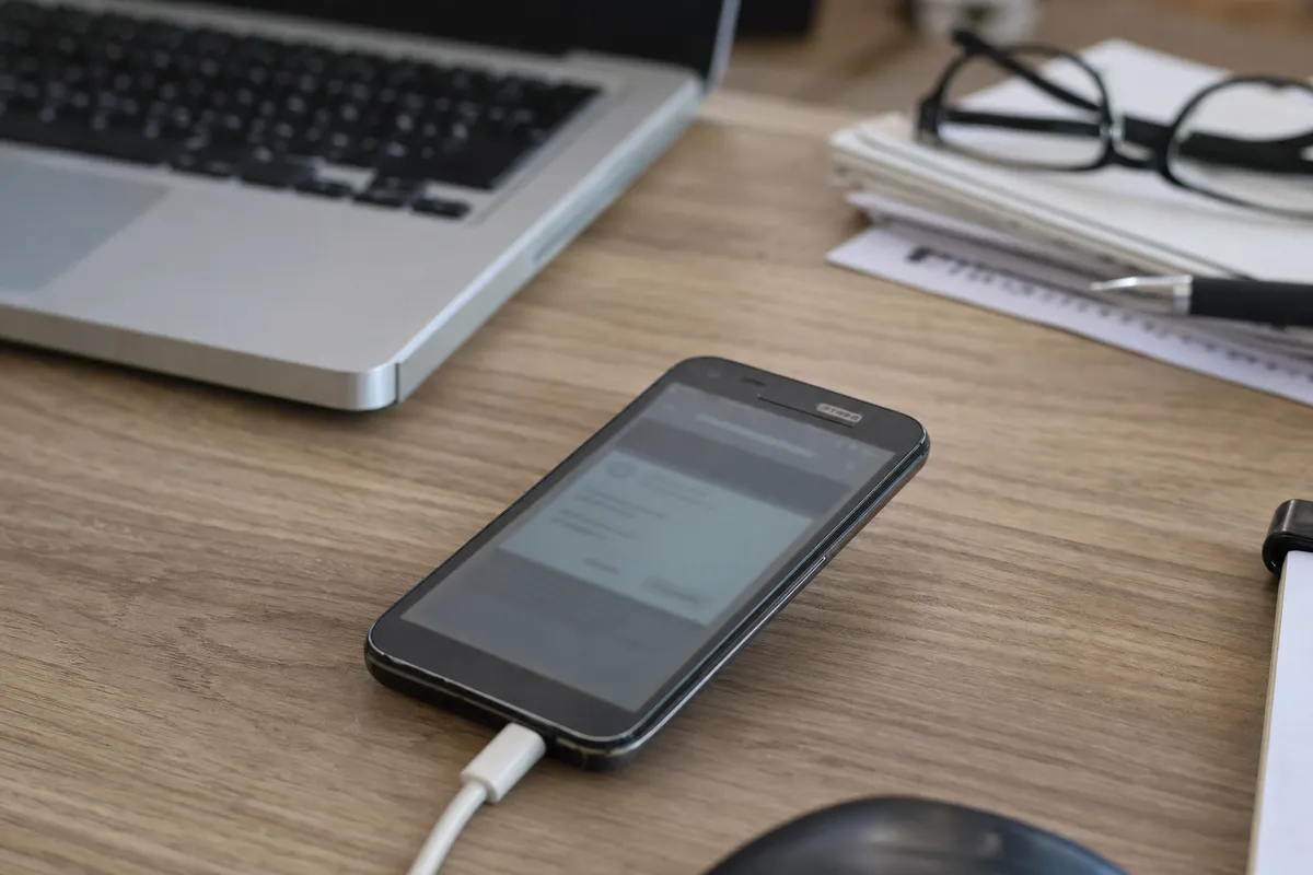 Vecchio smartphone Android collegato al cavo su una scrivania, con laptop sfocato e occhiali sullo sfondo