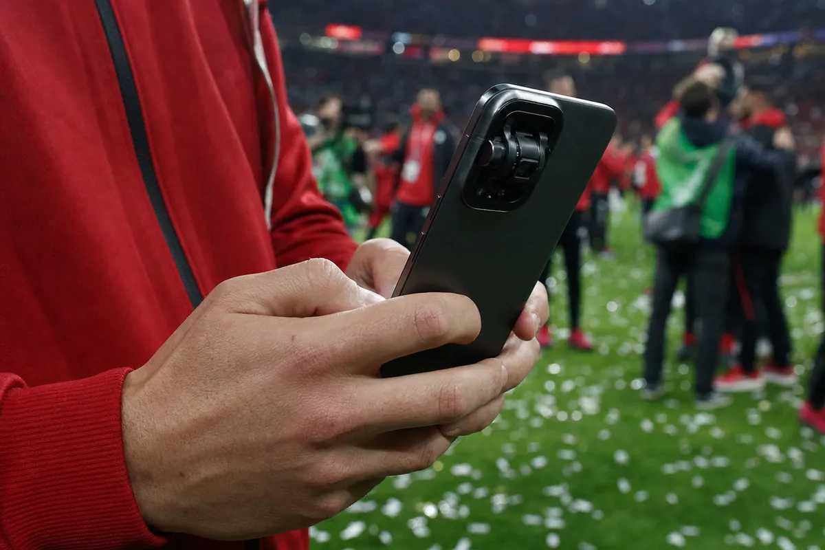 Mani di un giocatore con giacca rossa che regge uno smartphone scuro con grande modulo fotocamera, sul campo con coriandoli