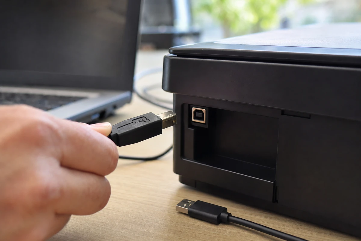Connettore USB Type-B vicino alla porta di una stampante, con estremità USB-A sul tavolo e laptop sullo sfondo