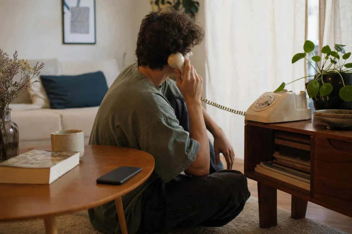 Giovane seduto in salotto usa un telefono fisso vintage con filo, mentre uno smartphone resta sul tavolino
