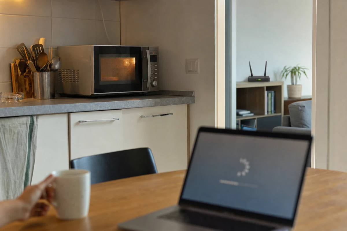Forno a microonde acceso in cucina, router Wi‑Fi su una mensola in un’altra stanza e laptop con schermata di caricamento