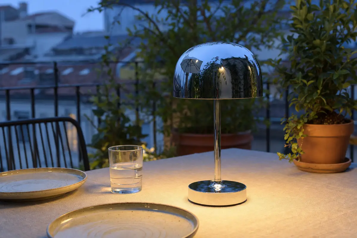 Lampada portatile cromata accesa su tavolo da terrazza con piatti, bicchiere d’acqua e piante al crepuscolo