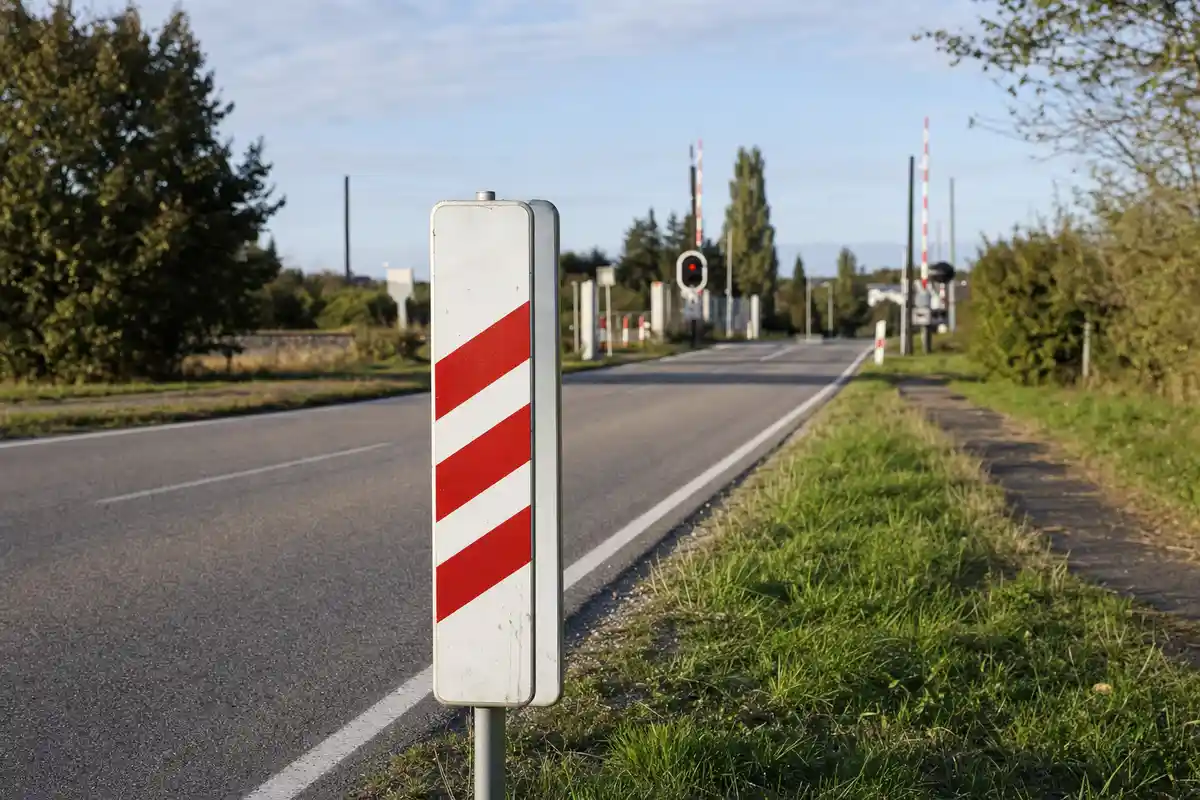 Panel rectangular blanco con tres franjas rojas en el arcén de una carretera rural, con paso a nivel al fondo