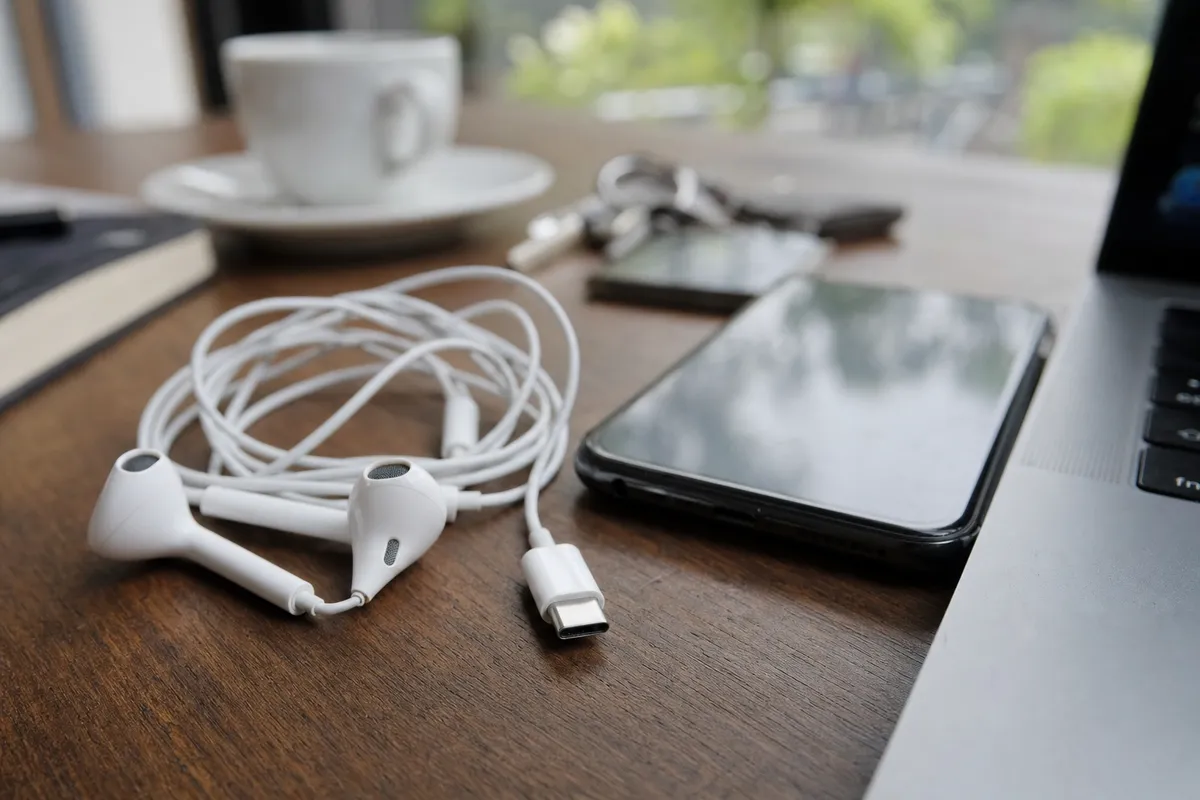 Auricolari bianchi con filo e connettore USB‑C su un tavolo, accanto a smartphone e bordo di un laptop