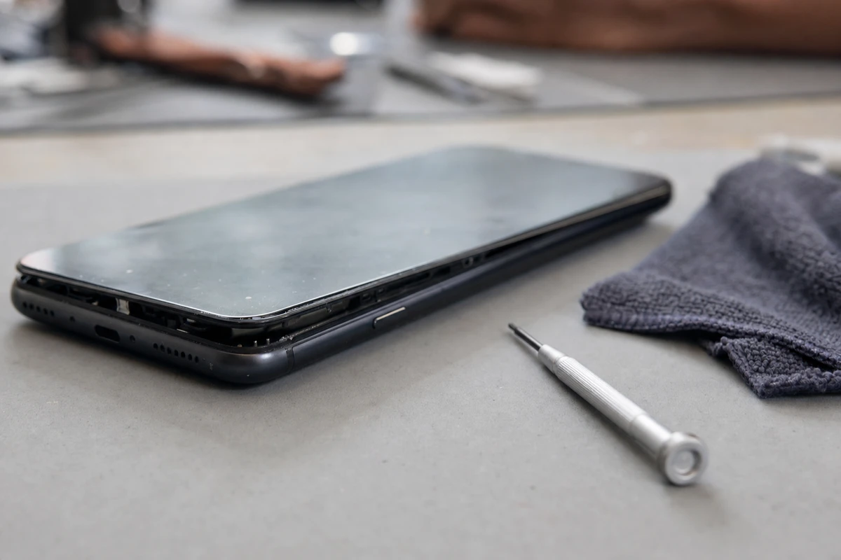 Smartphone con schermo sollevato dal telaio su un banco, con cacciavite di precisione e panno in microfibra