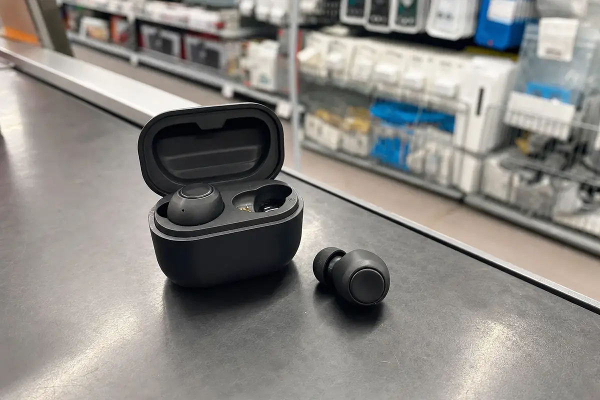 Auricolari wireless neri con custodia di ricarica aperta su un bancone, con scaffali di elettronica sfocati sullo sfondo