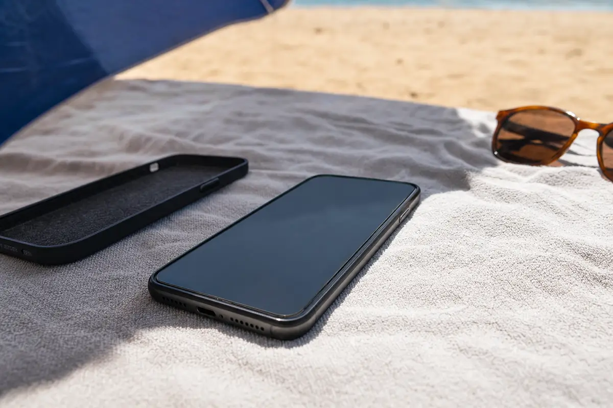 Smartphone su un asciugamano in spiaggia, con cover rimossa accanto e occhiali da sole sullo sfondo