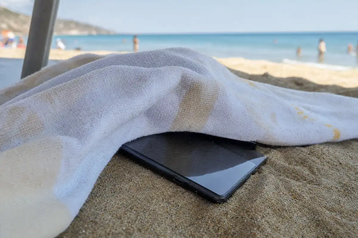 Smartphone nero semi coperto da un asciugamano sulla sabbia, con mare e bagnanti sfocati sullo sfondo