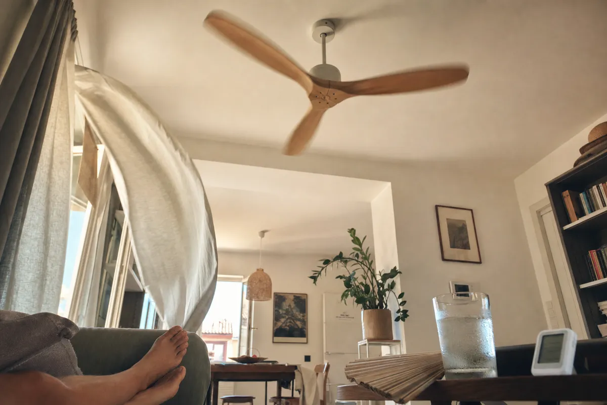 Ventilatore a soffitto in movimento in soggiorno, tenda che svolazza e bicchiere d’acqua su tavolino