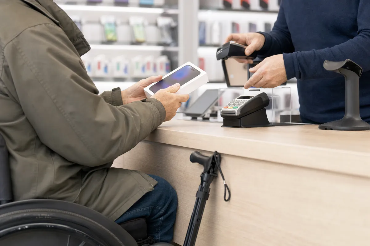 Cliente in sedia a rotelle acquista uno smartphone al bancone, con terminale di pagamento e addetto alla cassa