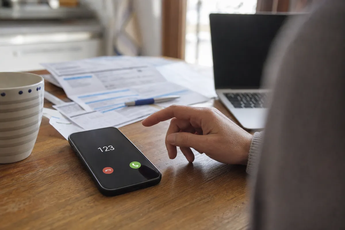 Smartphone su un tavolo con chiamata in arrivo dal numero breve 123 e mano esitante, con bollette e laptop sullo sfondo
