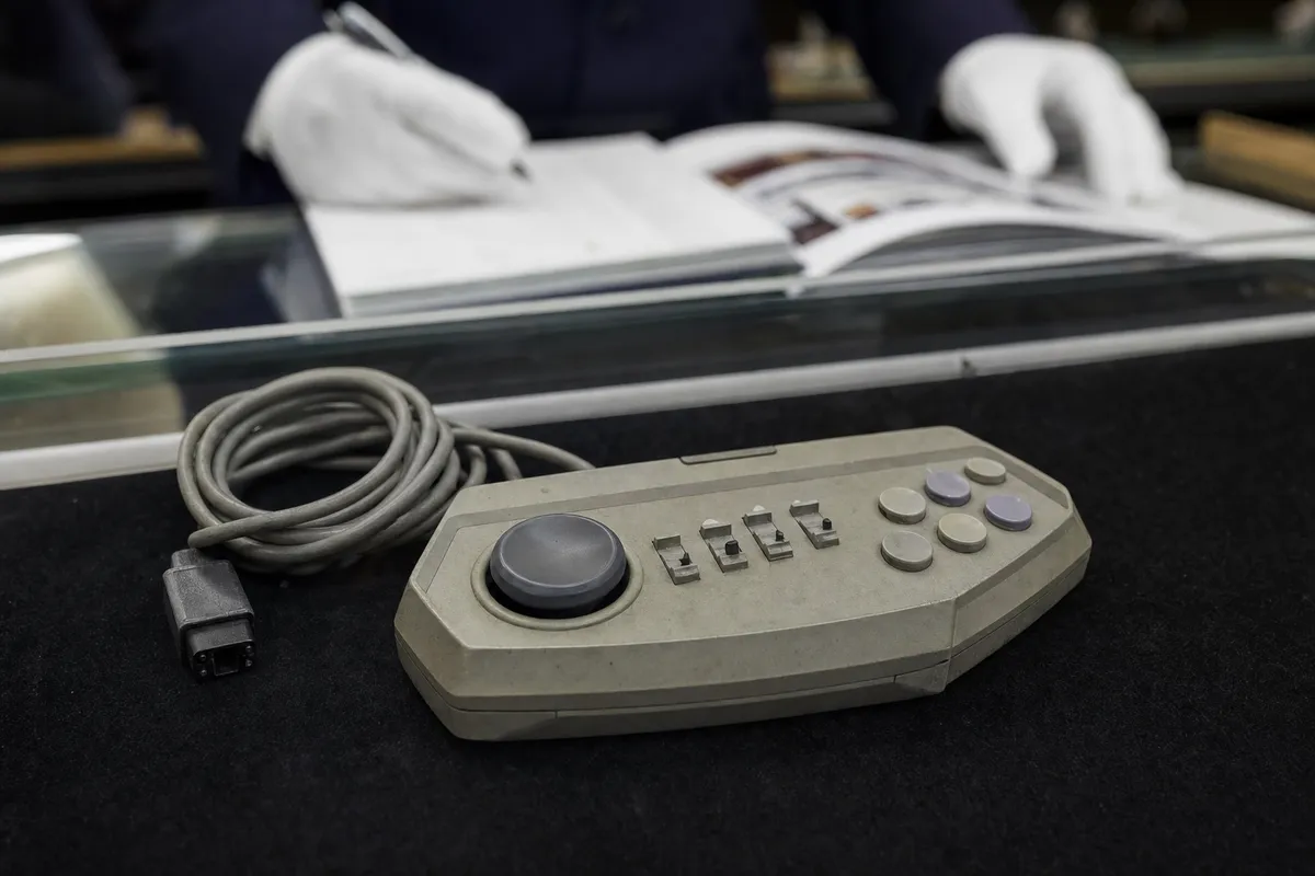 Controller vintage grigio con cavo arrotolato su panno nero, sullo sfondo mani con guanti bianchi e catalogo sfocato