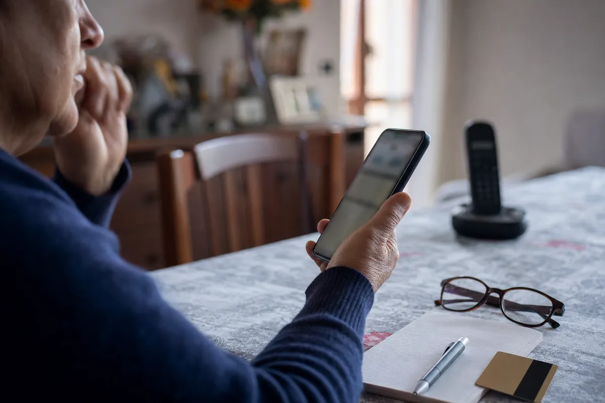 Persona al tavolo guarda uno smartphone durante una chiamata, con occhiali, taccuino, penna e carta sul piano