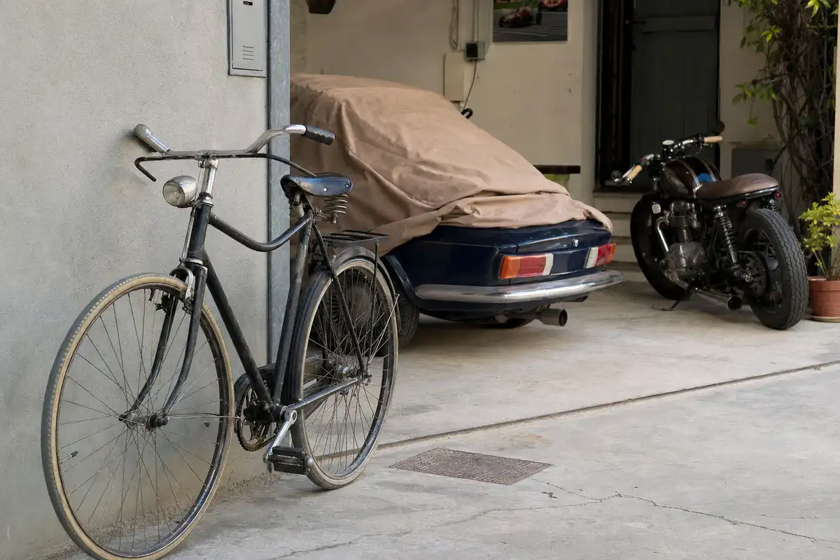 Bicicletta appoggiata al muro in un cortile, con auto d’epoca coperta e moto parcheggiata sullo sfondo