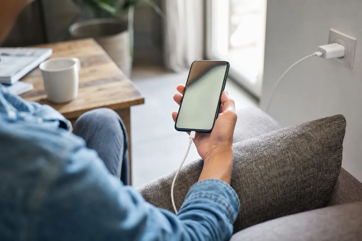 Mano che usa uno smartphone collegato al cavo di ricarica, seduti sul divano vicino a una presa a muro
