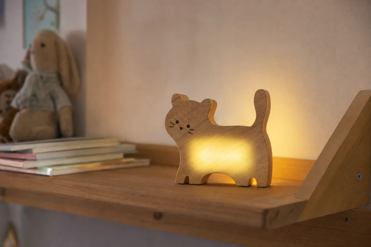 Lampada LED in legno a forma di gatto accesa su mensola in cameretta, con libri e peluche sullo sfondo