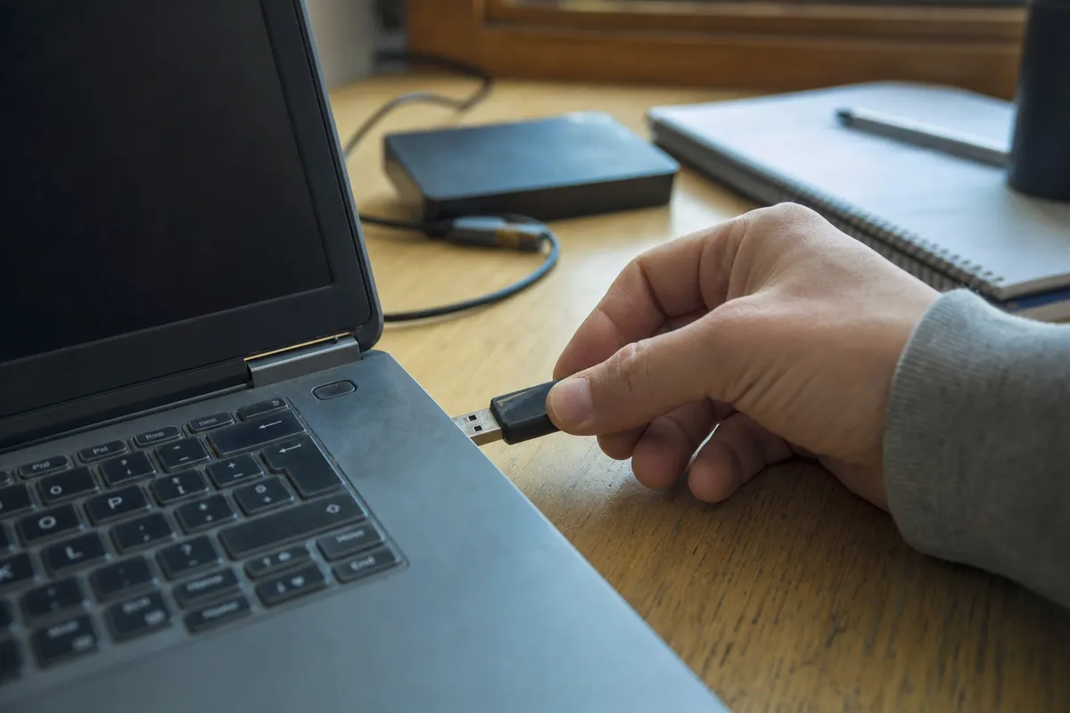 Mano che inserisce una chiavetta USB in un laptop su una scrivania, con hard disk esterno e quaderno sullo sfondo