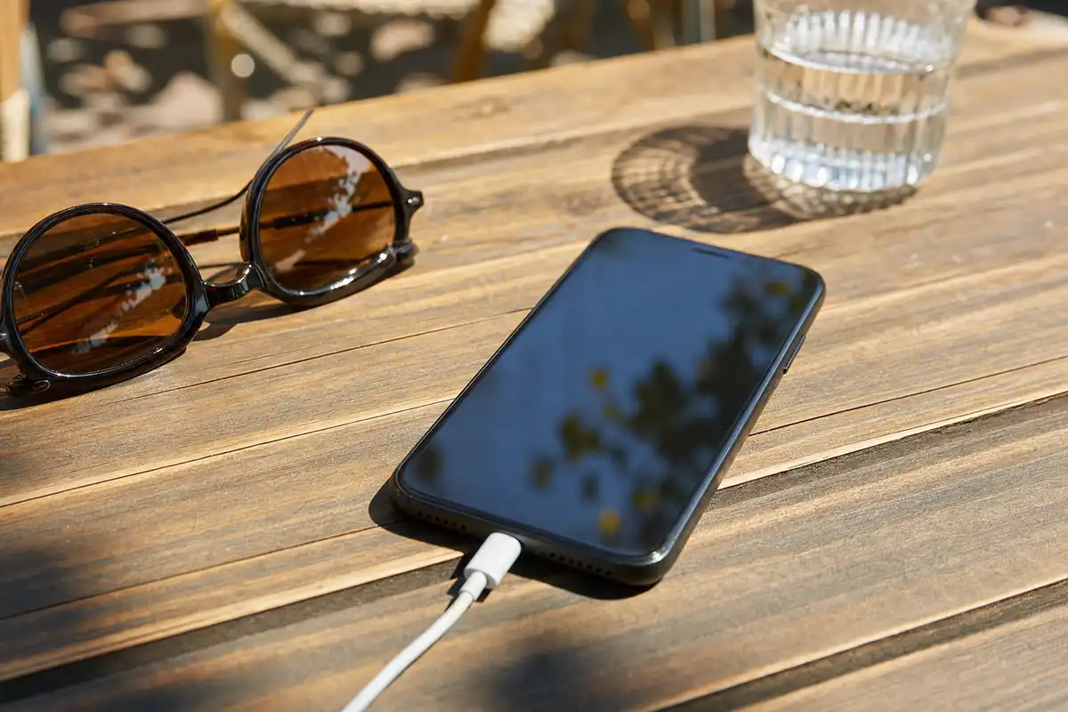 Smartphone con schermo spento in carica su un tavolo di legno al sole, con occhiali da sole e bicchiere d’acqua sullo sfondo