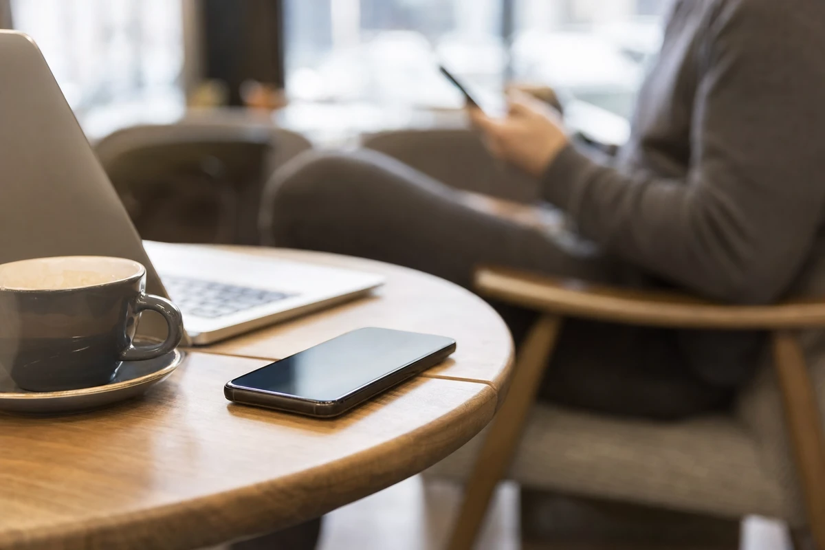 Smartphone appoggiato su un tavolino accanto a una tazza di caffè e a un laptop, con persona sfocata sullo sfondo