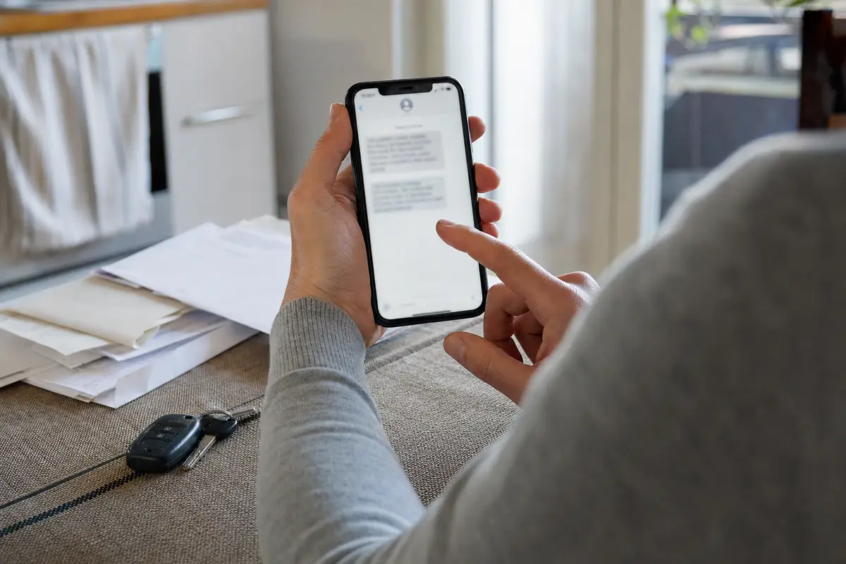 Mani che tengono uno smartphone con un messaggio sfocato, sopra un tavolo con chiavi dell’auto e pile di documenti