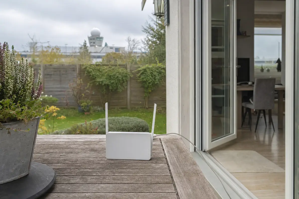 Router Wi‑Fi bianco su tavolo da patio con cavo verso casa, porta aperta sul giardino e radar in lontananza