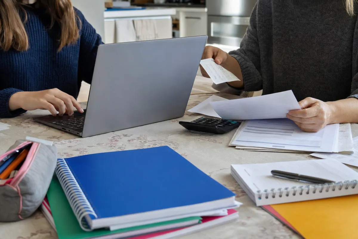 Laptop su un tavolo con quaderni e astuccio, mentre un adulto controlla documenti e uno studente usa la tastiera